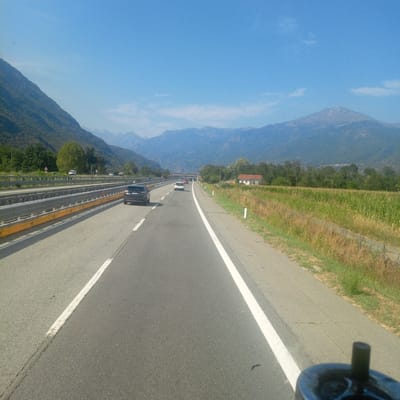 The image depicts a sunny day on a highway in Italy, likely near Calea. The highway is a multi-lane road with a clear lane marking and guardrails. Several cars are visible on the road, including a dark SUV in the foreground. The landscape is characterized by mountains in the background, with lush green fields and trees along the side of the road. A small building with a red roof is also visible in the distance. The sky is a clear blue with a few wispy clouds. The perspective suggests the photo was taken from a moving vehicle.