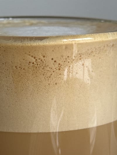 The image is a close-up of a foamy beverage in a glass mug, likely coffee or a similar drink. The foam is a light brown, creamy color with a texture of small bubbles. The beverage itself is a darker brown, visible through the glass. There are reflections on the surface of the glass, indicating ambient light. The background is a plain, light gray surface, possibly a table or wall. There are no people or discernible actions. The image is static and focuses solely on the beverage. The location of Гамово, Russia is not represented in the image.