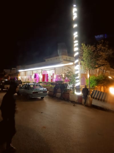 The image depicts a nighttime street scene in Boufakrane, Morocco. The central focus is a brightly illuminated commercial building, possibly a restaurant or cafe, featuring a light-colored facade with prominent window or archway sections. The exterior is adorned with warm white strip lighting, and the interior glows with vivid pink/magenta illumination. Green foliage from planters lines the base of the building. To the right, a tall, slender pole is wrapped with helical white strip lights, with trees visible around it and behind the building. In the upper right background, a blue neon sign displays inverted text.

The foreground consists of an asphalt road. A light blue sedan, bearing the Moroccan license plate "54296 | 1" and equipped with roof racks, is parked or slowly moving on the right side of the road, oriented away from the viewer. Part of a dark, obscured vehicle is visible on the far left. Concrete road barriers, one of which is striped blue and white, are positioned along the right side of the road.

Several individuals are present. A silhouetted, out-of-focus figure stands in the immediate foreground on the left. Two individuals are seated on a low wall or barrier in front of the commercial building. On the far right, an individual wearing a dark jacket and a light-colored cap stands beside a concrete barrier. Additional blurred figures are discernible near the obscured vehicle on the far left.