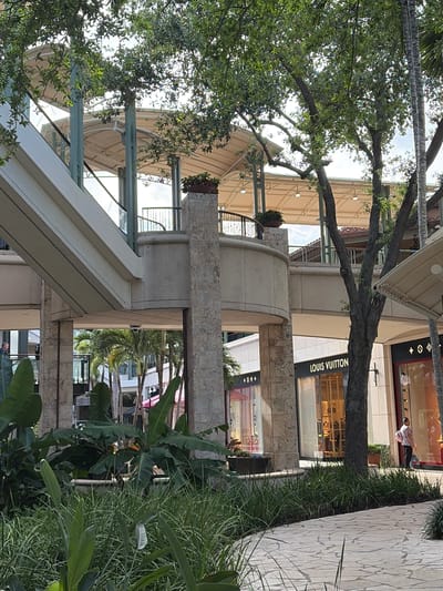 The image shows a horizontal shot taken in the city of Coral Gables, United States of America. The focus appears to be on a building or commercial structure, possibly a shopping center or retail location. Prominently visible is the Louis Vuitton store, suggesting a high-end shopping district. The architecture incorporates elements like a walkway, a cylindrical concrete structure, and a covered or shaded area, likely designed for pedestrians. The scene includes a variety of greenery, with plants and trees framing the built environment. In the upper right corner, an individual is seen walking near the Louis Vuitton store, adding a human element to the overall urban scene. The composition is angled upwards, capturing the building's height and the interplay between architecture and nature.