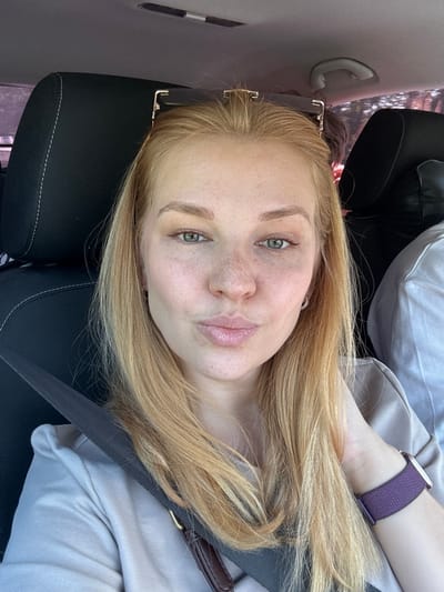 The image shows a close-up, selfie-style photograph taken inside a car. The subject is a young Caucasian woman with blonde hair, green eyes, and freckles. She is wearing a light-colored t-shirt and a purple watch on her left wrist. Her lips are pursed, giving the appearance of blowing a kiss. Her right hand is resting on her neck, and her left hand is out of frame. The background consists of car interior elements, including a black headrest with white stitching, a seatbelt, and part of another person wearing a white shirt. The lighting suggests it is daytime. The location is identified as Sosnovtsy, Russia, although this is not visually verifiable from the image itself.