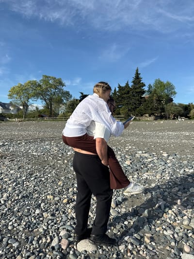 A couple is embracing on a pebble beach in Yuada-Gulrypsh, Abkhazia. One person wears a white shirt, brown pants, and white sneakers. The other wears a white shirt and black pants, with black shoes. The person facing the camera is taking a picture with their phone. The beach is covered in pebbles, and the background shows trees, buildings, and a blue sky.