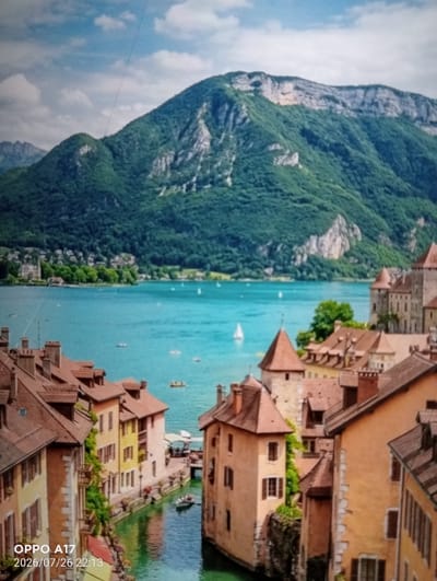 This is a photograph of a picturesque European town with canals and a large lake, nestled at the foot of a verdant mountain. The buildings lining the canals are old and colorful, with red tiled roofs and stone facades. Boats are visible in the canals and on the lake. Several sailboats are dotted across the turquoise water, with a distant shoreline featuring more buildings and trees. The mountain behind the town is covered in lush green foliage, with rocky outcrops visible. The sky is a clear blue with scattered white clouds. The scene evokes a sense of tranquility and old-world charm. The timestamp "OPPO A17 2026/07/26 22:13" is overlaid on the bottom left of the image. The location is not Құмөзек, Kazakhstan, but appears to be a European city like Annecy, France, or a similar alpine lake town.