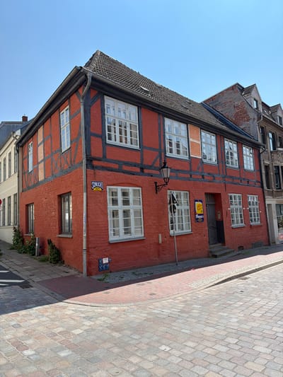 This image shows a corner of a historic brick building with dark timber framing, painted a vibrant red, under a clear blue sky. The building has multiple windows with white frames and is situated on a cobblestone street. To the left of the red building, a lighter colored building with vertical windows is visible. The building features a traditional German architectural style, common in the Hanseatic League cities like Wismar, Germany. A sign with the word "SUNLICHT" is attached to the side of the building. Another sign, with yellow and blue lettering, is hung above a door. The scene depicts a quiet street corner in what appears to be an old European town.