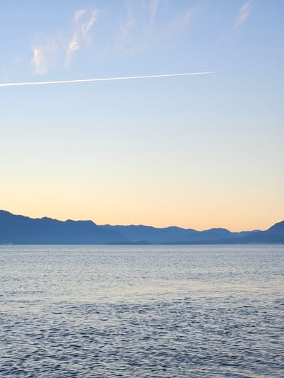 The image shows a tranquil scene in Villarrica, Chile. The right side of the frame is dominated by the reflective surface of Villarrica Lake, with gentle ripples creating a textural contrast. To the left of the lake, a silhouette of the Andes Mountains fades into the horizon. The sky transitions from a pale blue near the top left to a soft orange and yellow near the mountains, indicating either sunrise or sunset. A contrail from a passing aircraft cuts diagonally across the light blue portion of the sky.