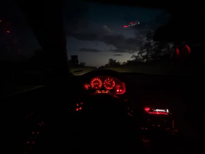 This image captures the interior of a car at night, looking out through the windshield. The dashboard is illuminated by red lights, displaying various gauges and a digital display that reads "CONSI 5.2 L/100". A car stereo is also visible, lit with red and white lights, showing the word "BUSINESS" on its display. Outside the car, the sky is dark with some clouds, and trees are visible on the sides of what appears to be a road. In the upper right of the frame, a bright, linear object with red lights is visible in the sky, possibly an aircraft. The overall scene suggests the car is in motion, driving through a rural or suburban area at dusk or nighttime. The location is identified as Aleksandrovo, Bulgaria.