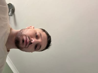 The image, rotated 90 degrees counter-clockwise, depicts an adult male from the chest up, positioned in the lower-left quadrant of the frame. He has light skin, short dark hair with faded sides, and a neatly trimmed dark beard and mustache. His dark eyes are directed towards the viewer, with a closed mouth and a neutral expression. A small dark spot is visible on his nasal septum. He wears a light-colored, possibly cream or pale yellow, long-sleeved garment, visible on his left shoulder and chest. The background consists of a plain, light-colored (off-white or light grey) wall. A metallic, silver-toned, ribbed wall-mounted spotlight fixture is visible in the upper-left quadrant, above and behind the male's head. A faint diagonal line, possibly a corner or molding, is visible in the bottom-left under his shoulder. The image depicts an indoor scene. No specific geographical location, such as Bretton, United Kingdom, is discernible from the visual content.