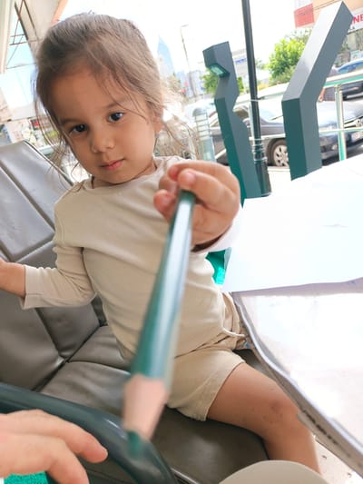 This image features a young child, likely a girl, sitting at a table outdoors. She is holding a long, green object, possibly a crayon or a pen, close to the camera, blurring the foreground. The child is wearing a light-colored, long-sleeved shirt and beige shorts. Her hair is pulled back loosely. Her expression is a gentle gaze towards the viewer.

The setting appears to be a public space, possibly a cafe or a park, indicated by the outdoor seating, a metal railing, and a street scene in the background. Cars and buildings are visible, suggesting an urban environment. Two dark, angular structures, possibly part of the seating area or a decorative element, are prominent in the mid-ground. The lighting suggests it is daytime, with bright sunlight. The overall mood is casual and candid.

There is visible text on a sign in the background, "NE SA", and a license plate on a car in the background reads "07 S." The location context provided is Ümraniye, Turkey.