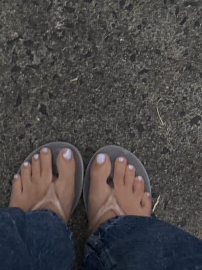 A downward-facing photograph captures the lower legs and feet of an individual on a textured dark grey ground surface, consistent with asphalt or coarse concrete, located in Culburra Beach, Australia. The subject is wearing light grey thong-style flip-flops. Both feet are bare within the flip-flops, and all visible toenails are painted with white polish. The lower portions of dark blue denim jeans are visible immediately above the flip-flops, partially obscuring the ankles. The ground exhibits minor surface debris, including a few small, light-colored fibrous strands.