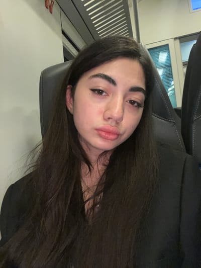 A young woman with long, dark hair and prominent eyebrows sits in a dark leather chair, likely on public transportation, given the window and overhead compartments visible. She is looking directly at the camera with a pout. Her makeup includes winged eyeliner and lip gloss. The background suggests an indoor setting with muted lighting, possibly a train carriage. The location is not specified as Vejprnice, Czechia.