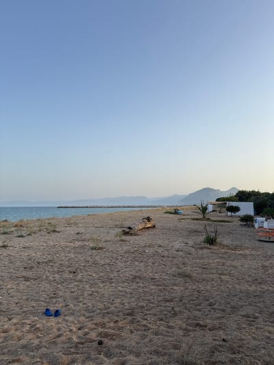 This image depicts a serene beach scene in Orosei, Italy. The foreground is dominated by a sandy beach with scattered dried vegetation and a large piece of driftwood. A pair of blue child's flip-flops rests on the sand in the lower left corner. In the mid-ground, a man-made structure, possibly a beach shack or cabana with a white awning, is visible, along with a small palm tree and some other sparse greenery. Further back, a breakwater made of large rocks extends into the tranquil, blue sea. The horizon is marked by a range of hazy, mountainous terrain. The sky is a clear, pale blue, transitioning to a lighter hue near the horizon, suggesting either early morning or late afternoon light. There are no people actively engaged in the scene, contributing to the peaceful ambiance. The overall impression is one of a quiet, undeveloped stretch of coastline.