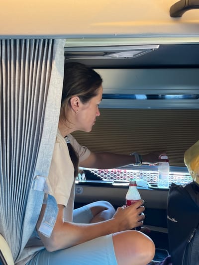 An interior shot shows a woman sitting inside a vehicle, possibly a camper van, looking to her right. She has dark hair pulled back and is wearing a light-colored t-shirt and blue shorts. Her right hand is reaching for a bottle of water on a table, while her left hand holds a red bottle with white liquid. She wears a watch on her left wrist and a ring on her right ring finger. The vehicle's interior features grey pleated curtains on the left and blinds on the right. The background appears to be a road or landscape visible through the window. The scene is lit by natural light coming from the window. The presence of the water bottle and the vehicle interior suggest travel. The image does not provide any specific visual cues to pinpoint the location to Ia Hru, Vietnam.