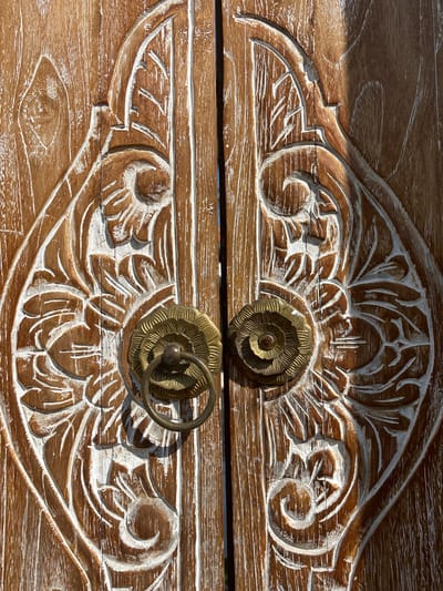This image shows two ornate wooden drawers with floral carvings. The wood has a light brown finish with white paint applied to highlight the carved details. Each drawer has a circular, brass-colored pull. The top pull is a solid flower design, while the bottom pull features a ring attached. The image was taken in Cemagi, Indonesia.