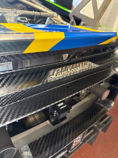The image presents a close-up view of the interior of what appears to be a Lamborghini race car. The dominant feature is a section of carbon fiber bodywork, prominently displaying the "Lamborghini" logo in gold lettering. Adjacent to this is a structure featuring a blue and yellow color scheme. Various mechanical components and wiring are visible in the background, contributing to the automotive context. The scene is likely located in Campagnano di Roma, Italy, given Lamborghini's connection to Italian automotive manufacturing. The composition suggests a detailed shot of the vehicle's construction or maintenance process.