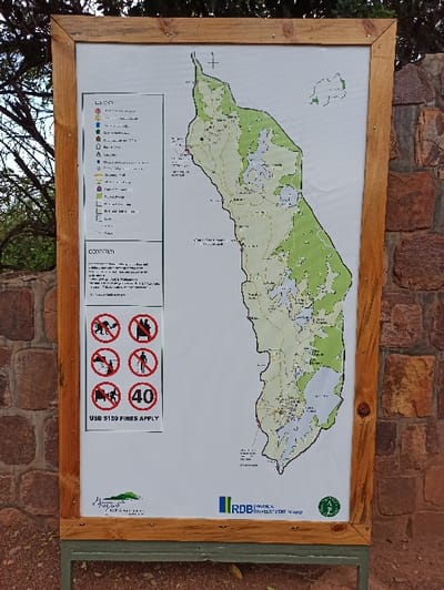 A framed informational sign displays a map of an elongated land area with varying shades of green, suggesting different vegetation types. Water bodies are depicted in white. Below the map is a legend identifying symbols for trails, points of interest, and other features. Prohibited activities are indicated by crossed-out icons, including cycling, motor vehicles, and hunting, with a speed limit of 40. The sign includes logos for RDB and Akagera Management Company, along with a warning about fines of USD $150 for violations.