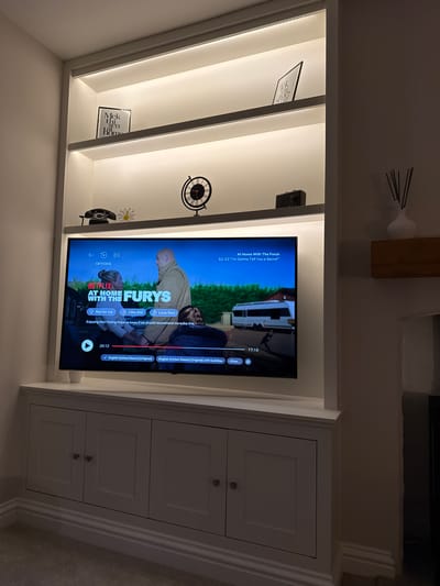 The image captures a bespoke, built-in entertainment unit in an indoor setting, likely a living room, in Harrogate, United Kingdom. The unit features white cabinetry with integrated lighting illuminating several shelving sections on the left, and three closed cabinets with simple knobs on the right.

At the center of the unit, a television is displaying the Netflix streaming service. The screen shows a scene from "At Home With The Furys", S2: E2, titled "I'm Gonna Tell You a Secret". The interface indicates a current playback time of 20 minutes and 12 seconds, with 17 minutes and 13 seconds remaining in the episode. Options for user interaction are visible, including "Not for me", "I like this", "Love this!", and a prompt asking "Enjoying this? Rating helps us know if we should recommend more like this." Subtitle options are also displayed, showing "English (United States) [Original]" selected, with a second "English (United States) [Original] with Subtitles" option.

The lit shelving on the left holds various decorative items. From top to bottom, these include a black speaker or camera-like device, a small framed picture displaying partially obscured text that reads "Mek thin home", a black decorative clock with Roman numerals, a small white daisy-shaped object, and a vintage-style black rotary telephone. Diffuser sticks in a white holder are visible on a wooden shelf above the entire unit. The integrated lighting creates a warm, inviting ambiance, suggesting the image was taken in the evening or in a room with controlled lighting.