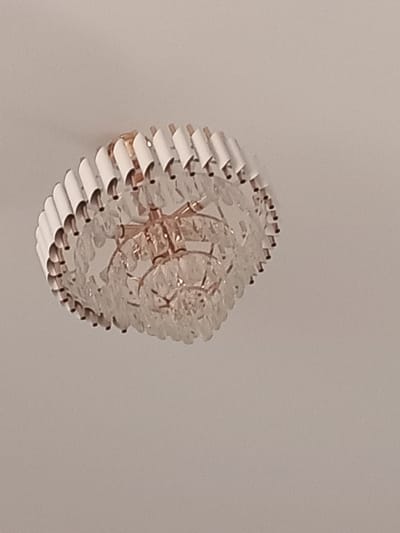 The image shows a close-up view of a decorative ceiling light fixture, possibly a chandelier, against a plain, light-colored background, likely a ceiling. The fixture is circular and appears to be made of glass or crystal elements suspended from a metal frame. The glass pieces are faceted and catch the light, creating a shimmering effect. The metal frame seems to be a warm, coppery or brass tone. The fixture has multiple layers of these glass elements, creating a textured and intricate appearance. Some of the glass pieces are elongated and have a ribbed or fluted texture, while others are more irregularly shaped. The outer edge of the fixture is lined with white, cylindrical elements. The lighting in the image is soft, suggesting it might be indoors during the day or with artificial light. There are no people, text, or strong location cues visible in the image itself, beyond the suggestion of an interior space. The context provided indicates the location is Navruz, Uzbekistan, which is associated with the Persian New Year celebration. However, the image itself does not depict any specific elements related to this holiday or location.