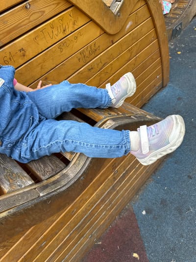 The image shows a child's legs and feet, wearing blue jeans and pastel-colored sneakers with white laces and Velcro straps. The child is lying on their back on a wooden bench, with their legs raised in the air. The bench is made of curved wooden planks, with some faint inscriptions on its surface. In the background, there's a blue and red speckled surface, possibly a playground surface. Another child, wearing a light blue shirt with a floral pattern, is visible lying on the bench to the left. The scene appears to be outdoors during the daytime, under clear weather conditions. The overall mood seems relaxed and playful. Some of the visible inscriptions on the bench include dates like "26.08.92", "6.4", and names or words like "Tuning", "Baby", and "WA".