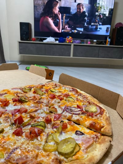 In Staraya, Russia, a close-up shot captures a pizza in a cardboard box placed on a light-colored floor. The pizza, topped with cheese, pickles, tomatoes, and red onions, is the primary focus. In the background, a television screen displays a scene with two people. To the left and right of the television are speakers. A gray media console with drawers sits below the television, holding various small objects, including a red ball and a white cap. The room's wall is painted a light color.
