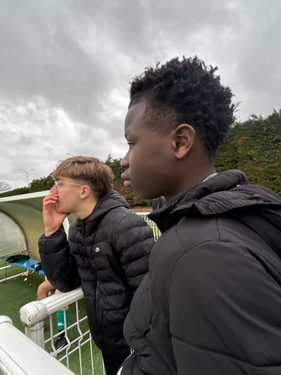 The image captures two young males and a partially visible third person on an overcast day at a sports complex in Montigny-lès-Metz, France. The scene is viewed from an elevated position overlooking an artificial turf sports field.
In the foreground, on the right, a young male with a dark complexion and short, dark, curly hair is shown in profile, facing right. He wears a black puffer jacket, and his gaze is directed intently outside the frame.
To the left and slightly behind the first individual, a second young male is visible in profile, facing left. He has light brown hair with a fringe, wears eyeglasses, and a stud earring in his left ear. He is dressed in a black puffer jacket with a white Lacoste crocodile logo on the left chest. His left hand is raised, with fingers near his mouth/chin, as he leans against a white metal railing that separates the viewing area from the field.
Behind the second male, part of a white-framed, semi-transparent dugout shelter is visible. Inside the dugout, a person's lower leg, clad in green shorts and black and white athletic cleats, is discernible on the artificial green turf field.
The background features dense green foliage, suggesting hedges or bushes on a raised bank, with faint outlines of utility poles visible further in the distance. The sky is heavily overcast with grey and white clouds. No direct interaction is observed between the two primary individuals.