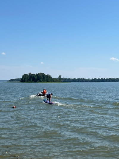 In the city of Borovoye, Russia, a bright, sunny day unfolds on a wide body of water. The sky above is a clear, vibrant blue, dotted with a few small, wispy clouds. The water itself is a deep, somewhat murky blue-green, with gentle ripples disturbing its surface, creating a slightly textured appearance. In the mid-ground, a small, inflatable boat with two individuals is navigating the water. One person, wearing an orange life vest, is at the helm, while the other is standing. Behind them, a person is actively engaged in what appears to be a water sport, possibly wakeboarding or surfing, as they are standing on a board and moving across the water, leaving a visible wake. In the foreground, to the left, a person's head is visible just above the water, indicating they are swimming or perhaps submerged. Across the water, a dense line of green trees forms the shoreline, with a small island covered in lush foliage situated further to the left. The overall scene depicts a recreational day on the water in a natural setting.