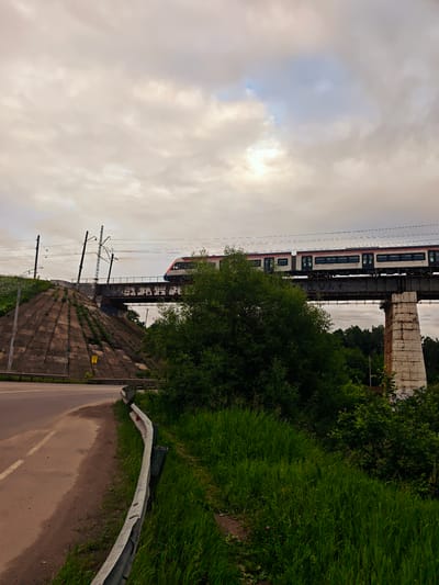An elevated train, primarily white with red and grey accents, is traveling across a railway bridge. The train has multiple carriages and dark tinted windows. The bridge itself appears to be constructed of metal girders and supported by a weathered stone pillar on the right side of the frame. Below the bridge, a large, dense green tree occupies a significant portion of the mid-ground.
To the left of the bridge, a concrete embankment slopes upwards, its surface covered in patchy green vegetation. A road, with a single white dashed line, curves away from the foreground towards the left. A metal guardrail runs along the edge of the road, separating it from a grassy verge. The sky above is overcast with diffused sunlight filtering through the clouds. The scene is set outdoors, with the environment suggesting a rural or semi-urban fringe. The image captures a snapshot of a train in motion, with the surrounding infrastructure and natural elements forming the backdrop. This specific view is located in Khotkovo, Russia, near the railway line.