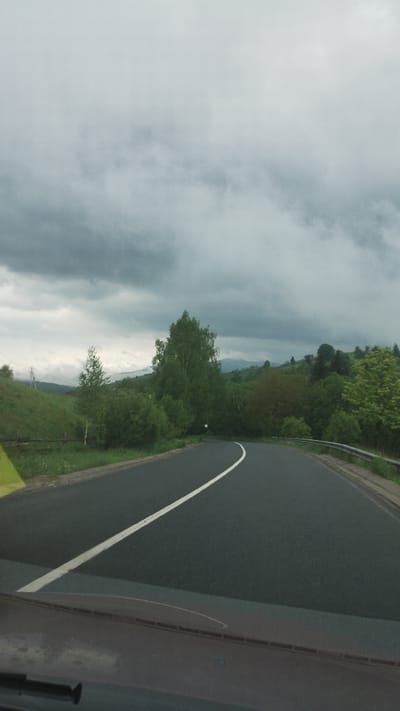 A gray asphalt road curves to the right of the frame, marked with a solid white line along its edge. Beyond the road, green vegetation suggests a hillside or valley. The sky is overcast with heavy, dark clouds. A portion of what appears to be a vehicle interior is visible in the lower right corner, including a section of a windshield. Without additional information, it's impossible to confirm the location in Lazeshchyna, Ukraine.