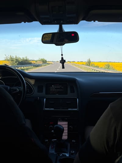 The image is taken from inside a car, looking forward through the windshield. The car is on a road that passes through a field of sunflowers. The sky is blue with some scattered clouds. On the dashboard, there is a navigation screen showing a map and route. A car accessory, a cross, is hanging from the rearview mirror. The road appears to be in a rural area, possibly outside the city of Buchach, Ukraine, given the presence of sunflower fields which are common in the region.