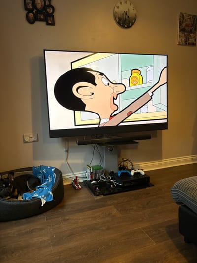 This image depicts a domestic interior, specifically a living area within a residence in the Forest of Dean, United Kingdom. The central focus is a large flat-screen television mounted on a wall, displaying an animated scene from the "Mr. Bean" series. On the screen, the cartoon character Mr. Bean is depicted with his mouth open, reaching into an open refrigerator or cupboard for a yellow container labeled "MUSTARD."

Below the television, a soundbar is positioned on a floating shelf. Beneath this, a tangle of cables and wires is visible. On the floor to the left of the TV stand, a black dog, possibly a French Bulldog or similar small breed, is curled up asleep in a black pet bed with a blue patterned blanket partially covering it. To the right of the pet bed and beneath the TV, various electronic devices are arranged on a low, black shelf. These include a PlayStation console, two gaming controllers (one blue, one white), and a stack of video game cases. A small red and white toy vehicle is also visible on the floor near the electronics.

The walls are painted a light, neutral color. On the wall above and to the left of the TV, a cluster of framed photographs is hung. To the right, a circular wall clock with a cityscape image is visible. Further right, a small framed picture is also affixed to the wall. The floor is covered with dark brown laminate wood flooring. A ribbed, grey upholstered cushion or part of a sofa is partially visible on the far right. A double electrical outlet is present on the wall to the left of the TV.