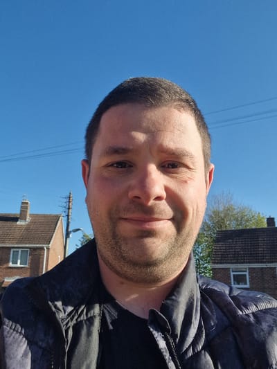This image is a brightly lit outdoor portrait of an adult male in Bearpark, United Kingdom. The man is positioned in the foreground, with his head slightly tilted and a gentle, friendly smile directed towards the camera. He has short, dark hair and stubble. He is wearing a dark-colored jacket, possibly padded or quilted, which is partially visible.
The background shows a residential scene, slightly out of focus. Several houses with brown tiled roofs and visible windows can be seen. Above these structures, a clear, vibrant blue sky dominates the upper portion of the frame, indicating bright and sunny weather. Thin power lines or telephone wires are subtly visible against the blue sky. The lighting suggests it is daytime, likely morning or afternoon, with direct sunlight illuminating the scene and creating soft shadows on the man's face. There is no specific activity or event depicted other than the portrait itself. No visible text is present in the image.