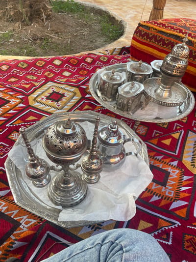 Two silver trays rest on a red patterned cloth. One holds a tea set, including a tall, ornate pot and several matching cups. The other contains a teapot, an incense burner, and two candle holders, arranged on a sheet of white paper. A person's leg, clad in denim, is partially visible to the right. The background reveals an outdoor setting with greenery and a structure possibly made of straw.