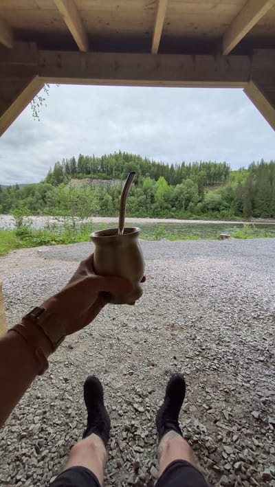 In Trofors, Norway, a person sits under a wooden shelter, holding a cup of mate with a bombilla. The person's legs are visible, wearing black socks and shorts. The background features a river flowing through a lush green forest, with a rocky path leading to the river. The sky is overcast.