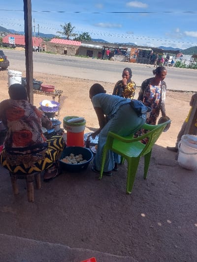 The scene depicts an active outdoor food preparation and sales area in Kimamba, Tanzania, under a bright, partly cloudy sky. The ground in the immediate foreground is compacted dirt or gravel, shaded by an unseen overhead structure supported by a dark wooden pillar on the far left.
Four individuals are present. On the far left, a person is seated on a low, three-legged wooden stool, viewed from the back. They wear a patterned dress featuring orange, red, black, and yellow floral motifs, with a yellow and black patterned fabric covering their legs. To their right, another individual is bent forward, facing away from the viewer, actively handling or preparing food items in containers on the ground. This person wears light blue clothing and a dark fanny pack around their waist. Adjacent to them is an upright green plastic chair.
Arranged on the ground near these individuals are several items: multiple silver cooking pots/containers, a purple bowl containing white, spherical objects, an orange and white insulated container, and a dark blue basin filled with numerous light-colored, spherical food items. A white bucket is visible on the far right.
Beyond the shaded area, a paved road stretches horizontally across the mid-ground. Two women stand near the road. The woman on the left wears a yellow and black patterned garment and has her hand near her mouth. The woman to her right wears a multi-patterned dress (light blue, brown, white, black) and holds a dark, possibly metallic, container in her left hand.
In the background, across the road, are various single-story buildings, some with red roofs, indicative of shops or residences. A dark colored vehicle is partially visible on the left side of the road. Utility poles and lines are visible overhead. Distant hills are present on the horizon under the blue sky.