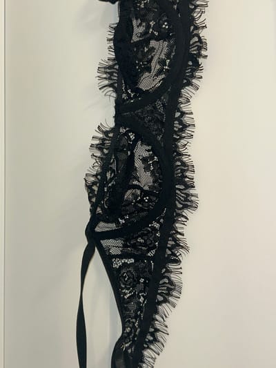 The image shows a black lace bra hanging against a white wall. The bra is detailed with intricate floral lace patterns and a frayed edge along the straps and cups. A thin strap extends downwards from the bra. The lighting is bright, casting subtle shadows on the wall behind the garment. The setting is indoors, with a plain, neutral background. No specific location within Lynnwood, United States of America, can be identified from the provided image.