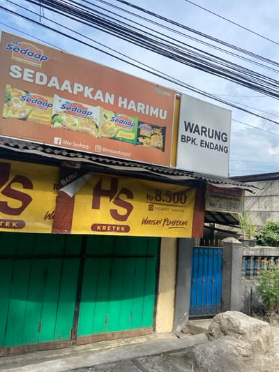 The image shows the exterior of a storefront located in Kecamatan Dayeuhkolot, Indonesia (latitude: -6.962, longitude: 107.614). The storefront is painted yellow with a large, orange banner above it. The banner advertises "Mie Sedapp" instant noodles. The sign reads "Sedaapkan Harimu" ("Make your day delicious"). The storefront has a green wooden door and a small, blue metal gate to the right of it. The gate is partially open. A large rock is visible on the ground in front of the store. The store is called "Warung Bpk. Endang" ("Mr. Endang's Store"). A second yellow banner is attached to the bottom of the store, advertising "HS Krettek" cigarettes for 8,500 rupiah. There is a third yellow banner on the right side of the store that advertises a service for "transport" and has a phone number, 0823-2418-161. Above the storefront, there are numerous power lines running overhead. The sky is mostly clear with only a few clouds visible.