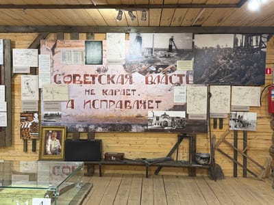 This is a medium shot, eye-level indoor photograph of a museum exhibit. The exhibit is displayed on a wooden wall with a wooden floor. The wall features a large mural with Russian text, a framed portrait of a man in a white suit, a black briefcase, and several historical photographs and maps. To the left of the mural, there is a glass display case containing historical items. To the right of the mural, a wooden bench displays a smaller brown briefcase, tools, and a pot. A fire extinguisher is visible on the far right side of the wall. The overall impression is one of a historical exhibit related to the Soviet era, possibly in a mining or industrial context given the presence of mining structures in the photographs and the tools on display. The location in Ust'-Kabyrza, Russia, is not explicitly stated in the image itself, but this context is provided.