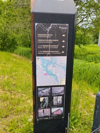 The image captures a black, weathered informational signpost positioned outdoors amidst vibrant green grass and foliage. The sign features multiple white panels on a black background, detailing local points of interest in Lithuanian and English, accompanied by a map and historical photographs.

The upper panel presents four points of interest with right-pointing arrows: "KABAMASIS TILTAS" (SUSPENSION BRIDGE), "PRIEKULĖS KLIOŠIŲ PARKAS" (PRIEKULĖ KLIOŠIAI PARK), "J. F. P. ŠRĖDERIO KAPAVIETĖ" (BURIAL SITE OF J. F. P. SCHRÖDER), and "J. F. P. ŠRĖDERIO SPAUSTUVĖ" (PRINTING HOUSE OF J. F. P. SCHRÖDER). Above this, a faded white outline of a region is labeled "KLAIPĖDOS APSKRITIS KLAIPĖDOS RAJONAS PRIEKULĖS SENIŪNIJA," indicating the sign's location within the Klaipėda County, Klaipėda District, Priekulė Eldership in Lithuania.

Below the main text, a blue and white map displays local waterways, roads, and settlements, including "Priekulė" and "Gropiškiai." A prominent red rectangle on the map, labeled "Jūs esate čia" (You are here), indicates the current location to be in or very near Gropiškiai, Lithuania.

The lower panel showcases four black and white photographs: an aerial view of a wooden suspension bridge over a river, a dense wooded area representing Priekulė Kliošiai Park, a fenced structure believed to be J. F. P. Schröder's burial site, and a rustic, single-story building identified as J. F. P. Schröder's printing house.

The sign itself exhibits wear, with some scratches on its surface and rust along its top edge. The surrounding environment consists of lush, untamed green grass in the foreground and a backdrop of verdant trees, suggesting a natural, rural or park-like setting under bright, possibly overcast, daylight conditions. No individuals are present in the scene.