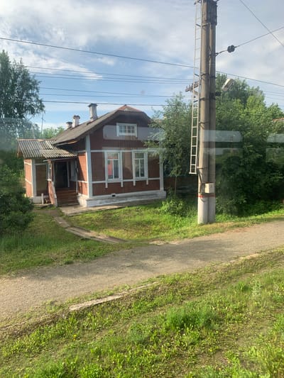 An exterior, eye-level, medium shot captures a wooden house in Berezovo, Russia, on a sunny day with a partly cloudy sky. The house, painted reddish-brown with white trim, features a gabled roof and multiple windows. A covered porch with stairs leads to the entrance. To the right of the house is a tall, concrete utility pole with a ladder attached and a street lamp. Lush green trees and grass surround the property, and a gravel path runs in front of the house. Power lines crisscross the sky above. The scene depicts a residential area in a Russian town.