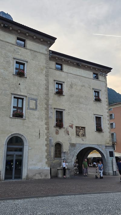 The image captures a view of a building in Riva del Garda, Italy. The building is old, with a weathered stone facade and arched doorways and windows. There are several people visible in the scene. In the foreground, a woman in black pants and a white shirt is holding a small white dog. To her right, a man in a green shirt and plaid shorts stands looking upwards. Further to the right, a woman in white pants and a blue top is using a walker. Behind them, through an archway, several tables and chairs are set up, suggesting an outdoor dining area. A motorcycle is parked near the archway. The sky is overcast, with a faint contrail visible. The street in front of the building is paved with cobblestones. The building appears to be part of a historic district in the city.