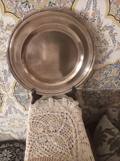 A top-down view captures a round, silver-colored platter resting on a surface composed of decorative tiles. The tiles exhibit a repetitive floral and geometric pattern in hues of white, yellow, blue, and orange. The platter is shiny, suggesting a polished metallic surface. To the right of the platter, a portion of a textured, possibly crocheted, off-white textile is visible, along with a section of dark fabric with a leaf-like pattern. The overall setting seems to be indoors, possibly a kitchen or dining area. The style of the tiles may indicate a location in Ksar-el-Kebir, Morocco, which is known for its traditional Moroccan tilework.