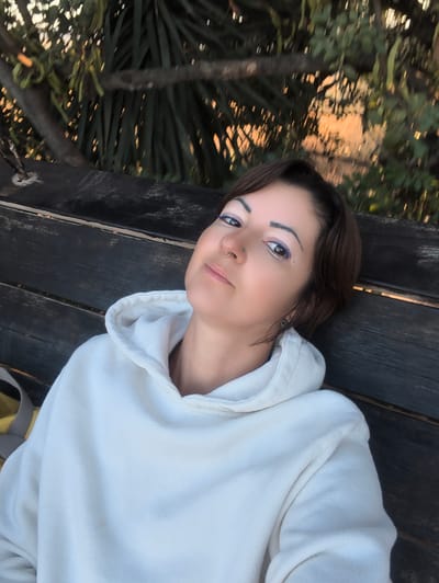 An adult female individual is shown in a close-up, upward-angled self-portrait, reclining on a dark wooden bench in Shaalvim, Israel. She has short, dark brown hair and is wearing a white hooded sweatshirt. Her face features prominent blue eyeliner on her upper lids, small gold glitter or stickers beneath her right eye, shaped eyebrows, and a gold hoop nose ring on her left nostril. A small stud is visible in her left ear. Her gaze is directed towards the camera. To her left, a portion of a yellow bag with a grey strap is visible. The background consists of out-of-focus dark green foliage and tree branches, with golden-hour sunlight filtering through, creating warm highlights and a dappled effect.