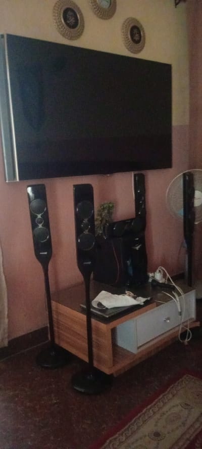 A dimly lit room with a large, flat-screen television mounted on a wall. Below the television, a wooden media console holds a sound system, wires, and a white cloth. Two tall, black speakers stand in front of the console, and another speaker is mounted on the wall to the right. A white standing fan is also visible on the right side of the image. Three circular wall decorations are positioned above the television. The floor is covered with a patterned rug. The overall scene suggests a home entertainment setup.