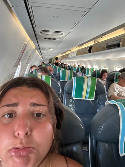 The image depicts the interior of a commercial aircraft cabin. The specific location within the city of Νέο Ρύσιο, Greece, cannot be determined from the visual evidence provided.

In the immediate foreground, a young adult female is partially visible, occupying the lower half of the frame. She has medium-brown hair, dark eyes, and a fair complexion with a reddish hue. Her lips are pursed in a pout, and a small silver hoop nose ring is present on her right nostril. She wears a dark, patterned strap top and a thin gold chain around her neck, looking directly at the camera.

Behind the foreground subject, the aircraft cabin extends rearward, showcasing multiple rows of dark-colored airline seats. Each seat is equipped with a headrest cover featuring vertical stripes in green, yellow, and light blue. The cabin is brightly illuminated by both natural light from windows and overhead artificial lighting. Numerous passengers are seated throughout the cabin. On the right side of the aisle, an older female passenger with white-grey hair and a light-colored top is visible, looking downwards. Further back on the left side of the aisle, a person with dark hair and a dark top is standing, reaching into an overhead compartment. Several overhead compartments are open on both sides of the cabin, revealing stowed luggage. The ceiling structure includes overhead service units with air vents and individual lights.