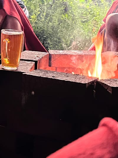 A close-up shot captures a glass of amber liquid on a dark surface, likely a table or counter, positioned to the left of a fire pit. The glass appears to contain a beverage with an ice cube and a straw. The fire pit itself is constructed from dark, rough-hewn bricks, with a vibrant orange and yellow flame emanating from its center, casting a warm glow. In the background, lush green foliage suggests an outdoor setting, possibly a park or garden, though its specific location within Bolshoy Istok, Russia, cannot be determined from the image alone. The presence of red fabric draped around the scene, potentially clothing or decorations, adds a splash of color to the composition. The overall impression is one of a relaxed, outdoor gathering, perhaps a barbecue or a casual get-together around a fire.