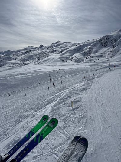 This image depicts a panoramic view of a snow-covered mountain ski slope within a ski resort in Saint-Martin-de-Belleville, France, viewed from an elevated perspective, likely a ski lift. The sky above is largely overcast with scattered clouds, though sunlight penetrates from the upper left, casting a subtle illumination on the scene.
In the foreground, the tips of two pairs of skis are prominently visible, indicating the viewpoint of an individual on a ski lift. The left pair of skis features a distinct green and blue design. The right pair of skis is mostly grey or silver and partially visible.
The midground and background showcase vast stretches of groomed snow, forming a wide ski run. Numerous individuals, appearing as small figures, are actively skiing down the slope, demonstrating various postures and apparent speeds. Visible along the edges and within the ski runs are red and yellow marker poles, delineating pathways and potential hazards. Several ski lift towers, supporting cables, are discernible along the mountain contours in the background and midground, suggesting an extensive lift system.
The distant landscape features undulating, snow-covered mountain ranges and peaks, some exhibiting exposed rock formations and sparse clusters of evergreen trees. The overall environment is one of an active winter sports area under partially bright conditions.