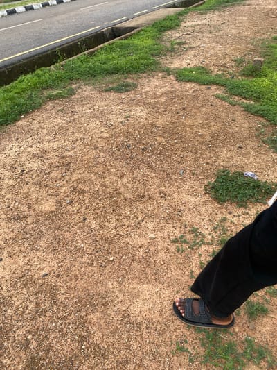 The image captures a close-up, slightly tilted view of a person's lower legs and feet, wearing black pants and black sandals. The setting appears to be outdoors, with a dirt and gravel ground in the foreground, interspersed with patches of green grass. To the left of the dirt area, a paved road with yellow and white markings is visible, bordered by a drainage channel filled with vegetation.
The lighting suggests it is daytime, possibly late afternoon given the angle of the light and the shadows cast. The weather appears to be clear. The scene evokes a casual, everyday moment, with no specific event or strong emotions conveyed. The location context is Nigeria, and the specific details of the road and vegetation hint at an urban or semi-urban environment. There is no visible text in the image.