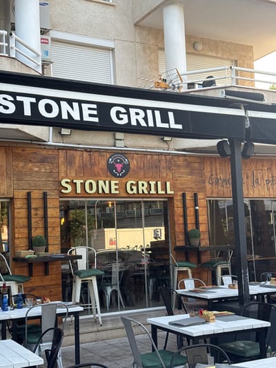 A vertical shot shows the exterior of a restaurant called "STONE GRILL". The restaurant has a rustic facade made of wood panels and a prominent black sign with white lettering that reads "STONE GRILL". The sign is angled and runs along the side of the building. To the right of the sign, another sign with golden letters reads "STONE GRILL" vertically. Below this, a circular logo features a bull's head with musical notes around it. Further right, outdoor seating is visible with white tables and chairs, some with green cushions. Bottles of condiments, including Heinz ketchup, are on the tables. Plants in woven baskets are placed on shelves on the wooden wall. The scene appears to be daytime, with sunlight reflecting off the building's windows. The location is Pla Parcial Platja, Spain, as indicated by the prompt. There are no people visible in the shot.