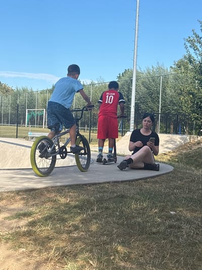 In Strombeek-Bever, Belgium, a skatepark scene unfolds under a clear blue sky. A young boy on a BMX bike with bright yellow tires is positioned in the center, his legs bent as he pedals. To his right, another boy in a red "BELGIUM 10" jersey stands on a scooter. A young woman sits on the concrete edge of the skatepark, looking at her phone. The environment is a typical outdoor skatepark with concrete ramps and surrounding grassy areas, bordered by a fence and trees.