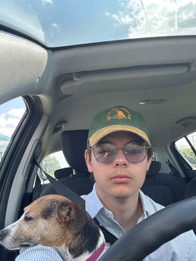 The image shows a young man wearing a green baseball cap with a gold emblem, round-rimmed glasses, and a blue and white striped shirt. He is sitting in the driver's seat of a car. There is a small, brown and white dog, wearing a pink collar, lying on the passenger seat. The image is likely taken from the passenger side of the car. The man is looking at the camera, with a neutral expression. The man and dog are in a car. The car's interior is visible. The image was taken in Pocklington, United Kingdom, a market town and civil parish in the East Riding of Yorkshire, England.