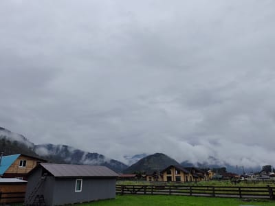 The image depicts a rural landscape under a cloudy sky, likely in Anos, Russia, given the context. In the foreground, a gray, rectangular building with a sloped, dark metal roof is prominently visible on the left. A wooden ladder is leaning against its side, leading towards the roof. To its left and slightly behind is a smaller wooden structure with a corrugated metal roof. Further left, a house with a blue metal roof and wooden walls can be seen.
Dominating the mid-ground is a wooden fence, partially obscured by tall green grass. Beyond the fence, a row of modern, wooden houses with dark roofs and large windows are situated, suggesting a residential development. These houses have a contemporary architectural style.
In the background, rolling hills and mountains are shrouded in low-lying clouds and mist, creating a dramatic and somewhat atmospheric effect. Patches of green vegetation are visible on the slopes of the hills. The sky is overcast with thick, gray clouds, indicating a likely overcast or potentially rainy day. The scene is devoid of any people or animals. The overall impression is one of a peaceful, if somewhat somber, rural settlement nestled amongst natural scenery.